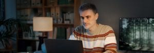 ストライプのジャンパーを着た男性が机に座り、目の前のノートパソコンを見ている。